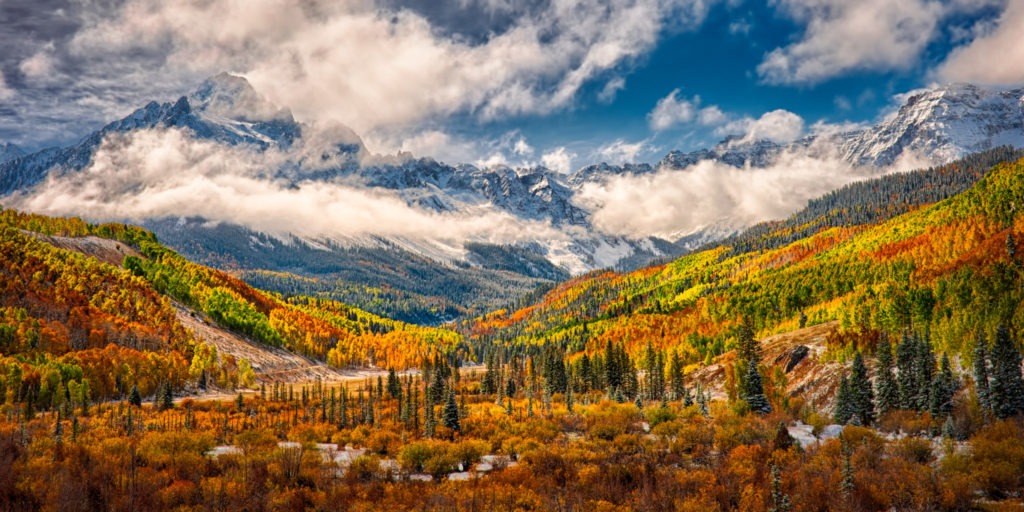 Colorful aspens and russet oaks treat the eye along CR 7 near Ridgway, Colorado, with the snow-capped mountains of the Sneffels Wilderness in the distance under retreating storm clouds.