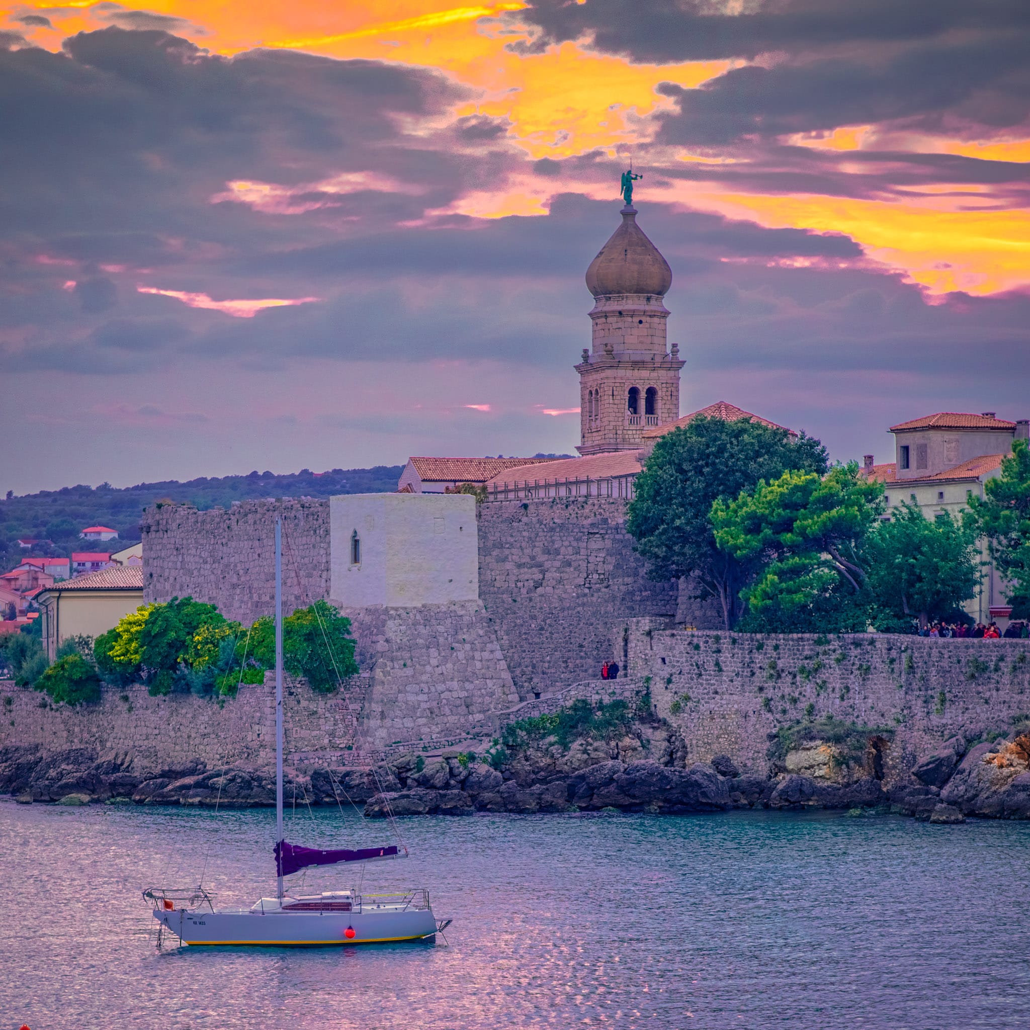 A sail boat sits at anchor near the medieval walls of Krk, Croatia, with Krk Cathedral in the distance.