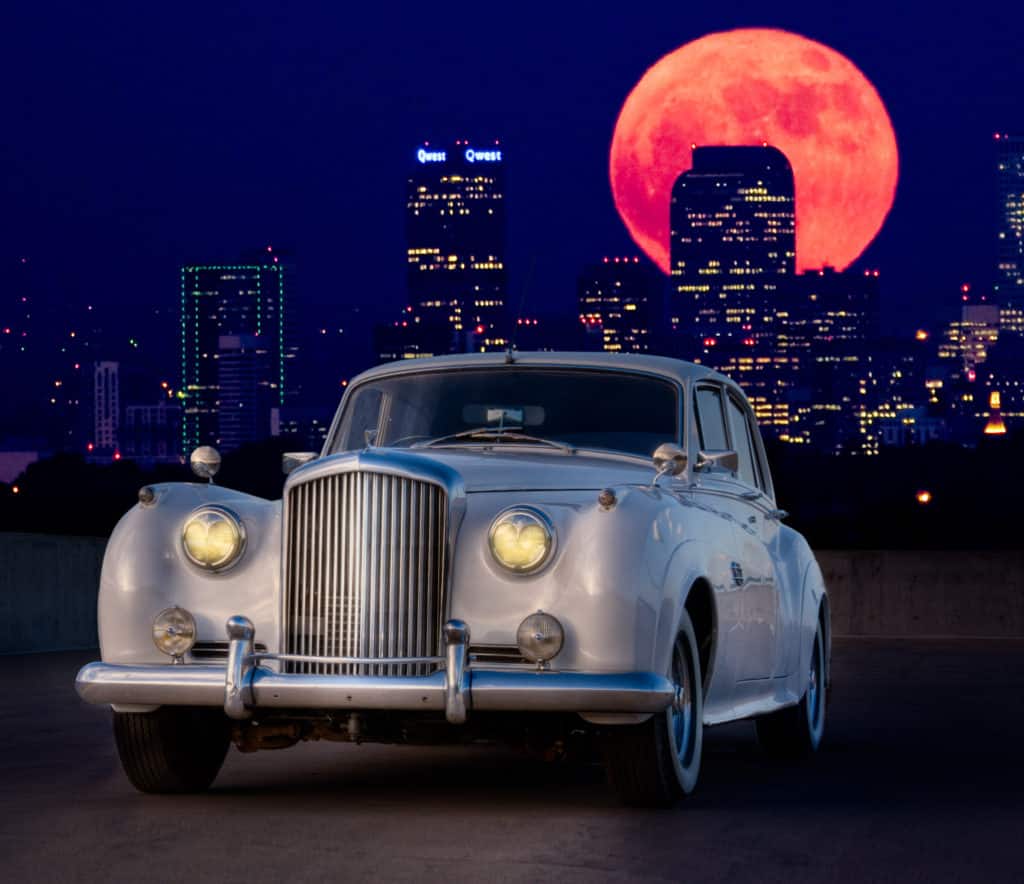 Photo of a 1959 Bentley S1 taken at night with a red moon and cityscape in the distance.