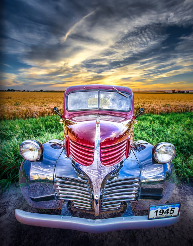 Front view of a 1945 Dodge Pickup Truck with red body and black fenders, sitting near a wheat field at sunset.