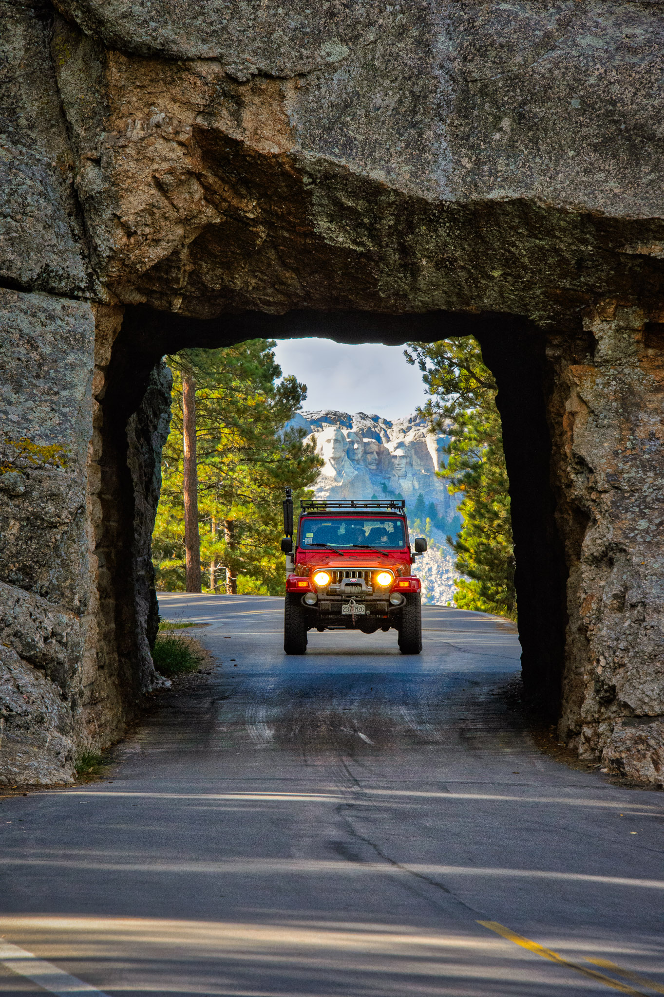 A red Jeep Wrangler enters a tunnel on the Iron Mountain Highway with Mount Rushmore visible in the distance.