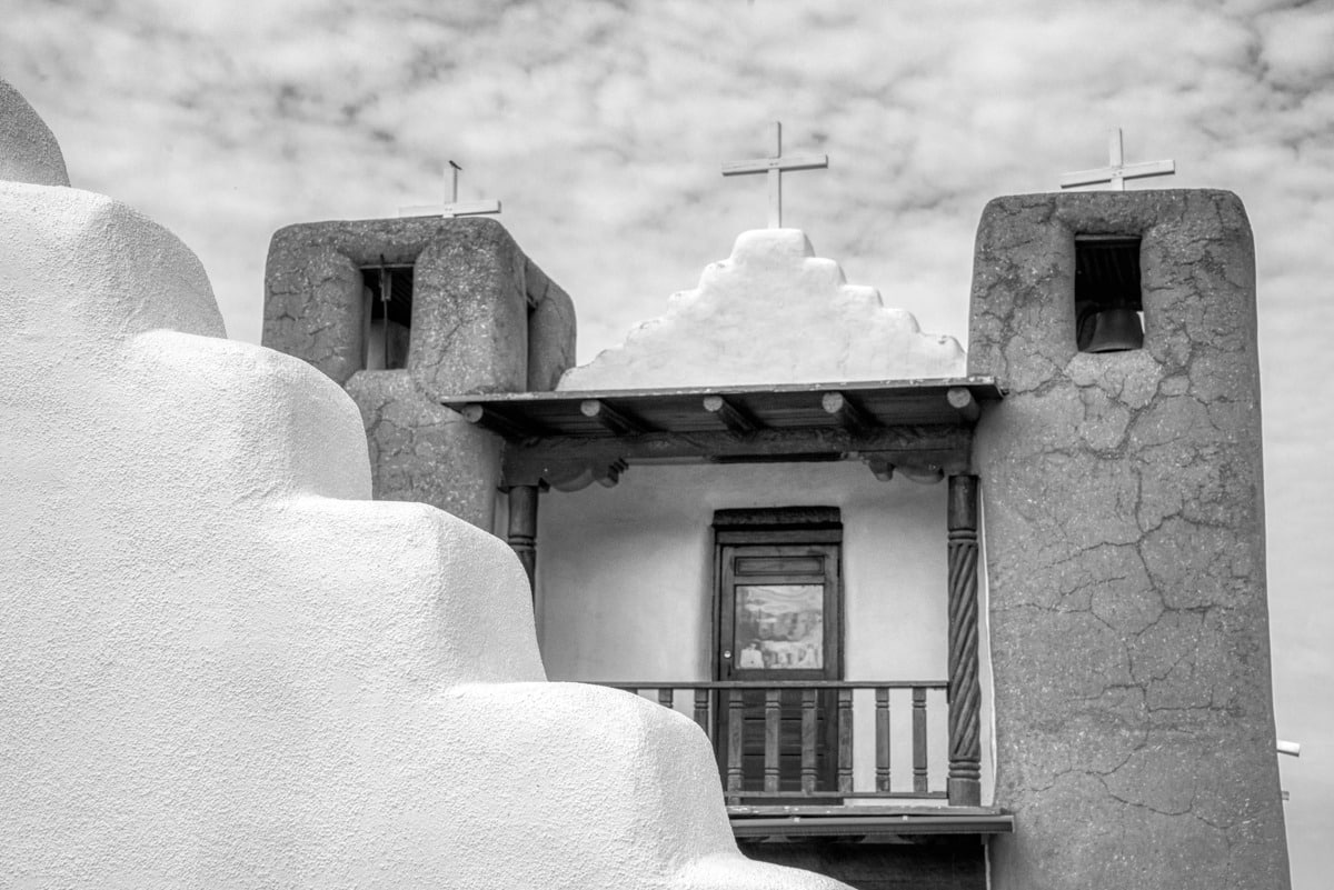 A black-and-white view of the balcony of St. Jerome Catholic Chapel with a portion of the arched gateway in front. The chapel is located in the heart of Taos Pueblo, New Mexico.