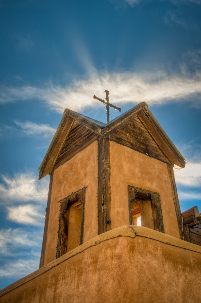 A view looking up at one of Santuario de Chimayo's bell towers with a cross at the tope. The cross is wrapped in mini Christmas lights. The santuario is located in Chimayo, New Mexico.