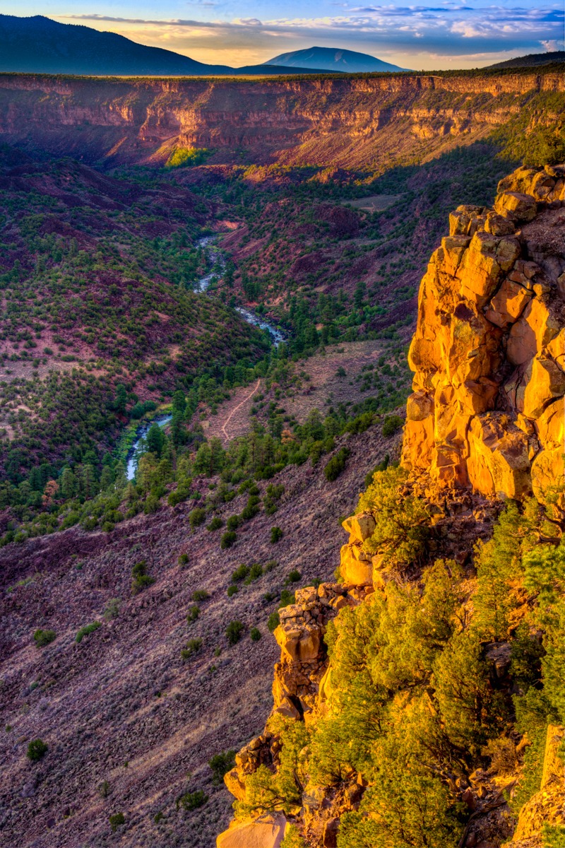 The cliffs and hills along the Rio Grande gorge in Wild Rivers Recreation Area are tinged orange and yellow by the setting sun.