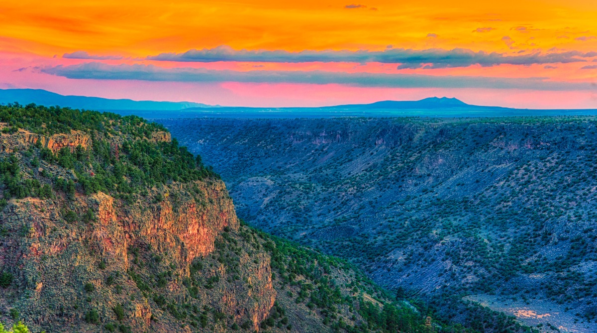 The cliffs and hills along the Rio Grande gorge in Wild Rivers Recreation Area are tinged orange and yellow by the setting sun.