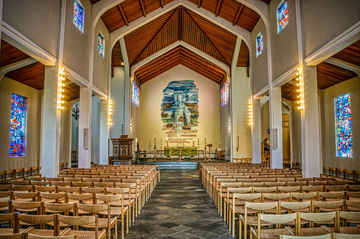 This is the interior view of Skálholt Church that features a vaulted ceiling, stained-glass windows, and mosaic altarpiece.