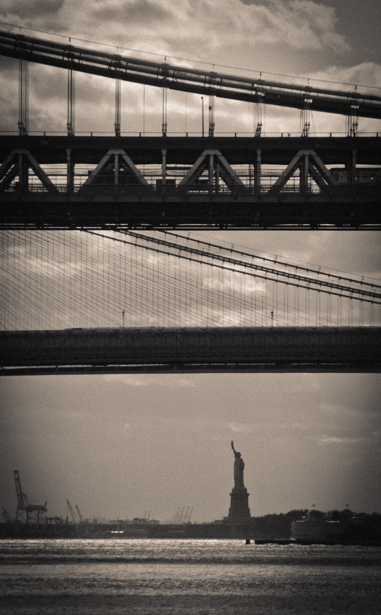 This foreshortened image shows the Statue of Liberty beyond the Brooklyn Bridge and the Manhattan Bridge in New York City.