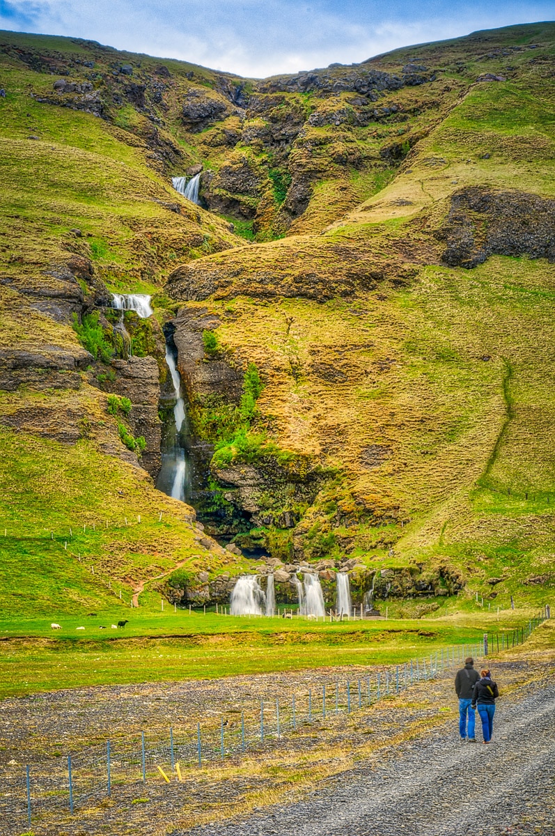 A couple walk down a side road toward a cascading waterfall called Merkjárfoss in southwestern Iceland.