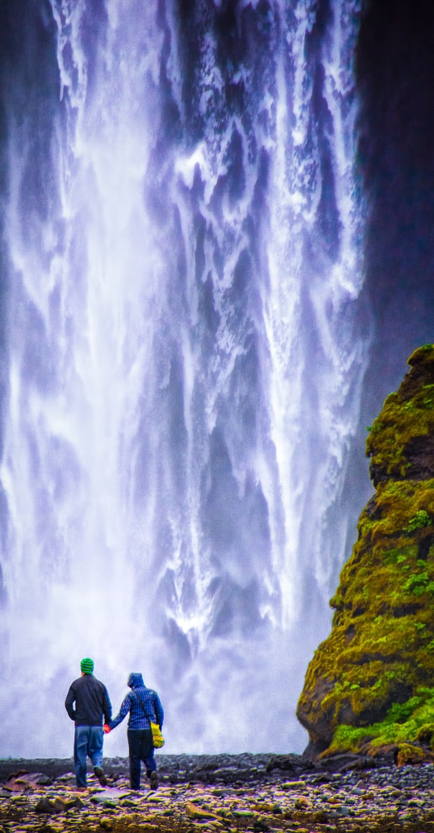 A couple are standing at the base of Skógafoss, looking at the base of the waterfall.