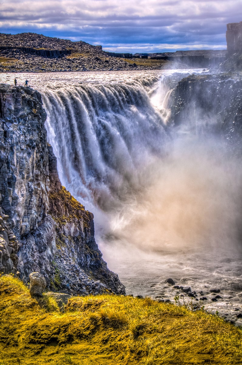 Two people stand near the edge of Dettifoss, the most powerful waterfall in Europe. It is located just south of Jökulárgljúfur National Park in Iceland.